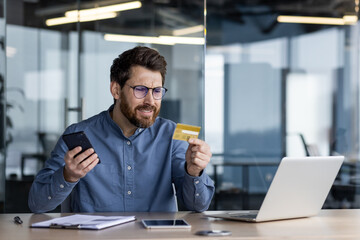 Worried young man sitting in the office at the desk, holding the phone in his hand and looking...