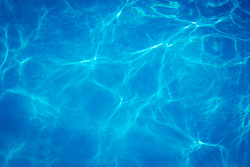 Clear blue swimming pool water surface with sunlight reflections creating abstract patterns and ripples, evoking a refreshing and tranquil summer vibe.
