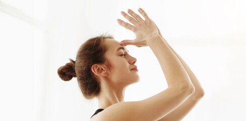 Fototapeta premium A serene young woman with her hair tied up in a bun is performing a yoga pose with a delicate hand gesture, her face exuding calmness. Sunlight gently illuminates her, closeup