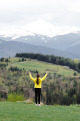 Girl in yellow jacket stands on hilltop with mountains in background