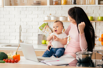 A mother and baby share a joyful moment in a modern kitchen. The baby sits on the counter while the mother looks at a laptop, both smiling and engaged.