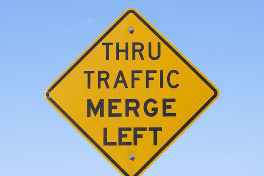 THRU TRAFFIC MERGE LEFT signage isolated against a blue sky background.