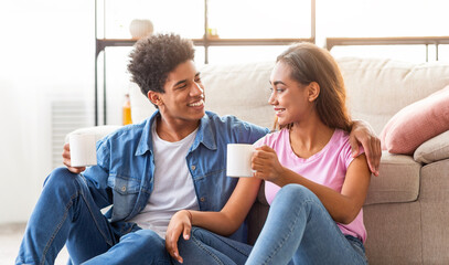 Black teen couple sitting on the floor in their living room. They are both smiling and holding coffee mugs, enjoying each others company. They are both looking at each other with affection