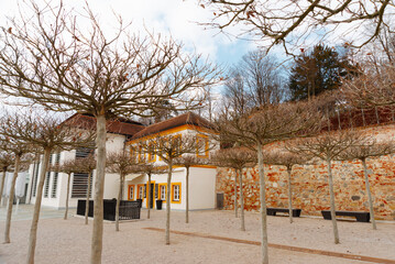 A serene courtyard with leafless trees and an ancient edifice in the backdrop, set against a cloudy sky