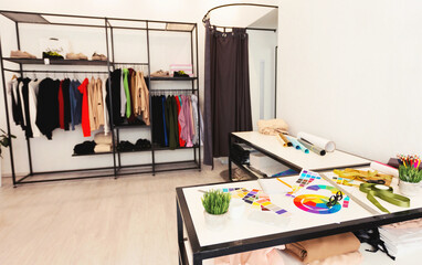 A room cluttered with various clothing items strewn across the floor and hanging on racks. A table in the center is piled high with more clothes, creating a chaotic and untidy environment.