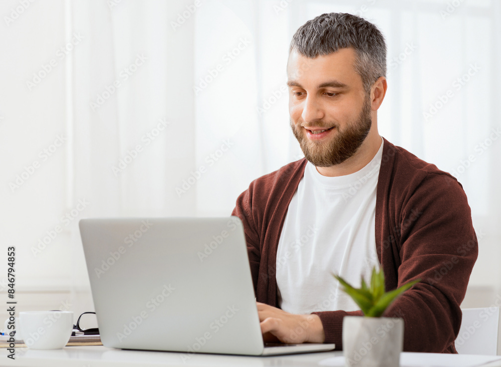 Poster A man is seated in front of a laptop computer, focused on the screen with a serious expression. He is typing on the keyboard and appears to be working or studying intently.