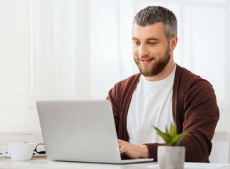 A man is seated in front of a laptop computer, focused on the screen with a serious expression. He is typing on the keyboard and appears to be working or studying intently.