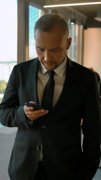 Portrait of a attractive businessman standing near window and writing sms. He look very smartand serious, slow motion. Slider camera movement. Vertical shot.