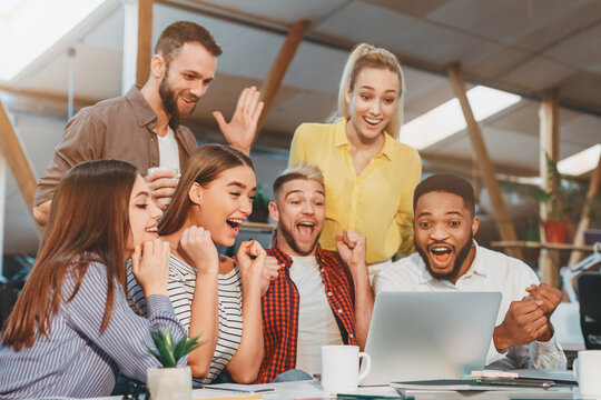 A diverse group of young professionals cheer and celebrate after achieving a milestone while gathered around a laptop in a contemporary office setting.