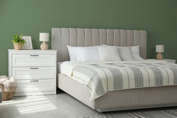 Comfortable bed and chest of drawers near sage green wall in room. Interior design