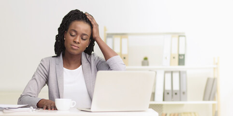 African American businesswoman suffering from migraine at office setting, sitting at desk in front of laptop with closed eyes, touching her head, copy space