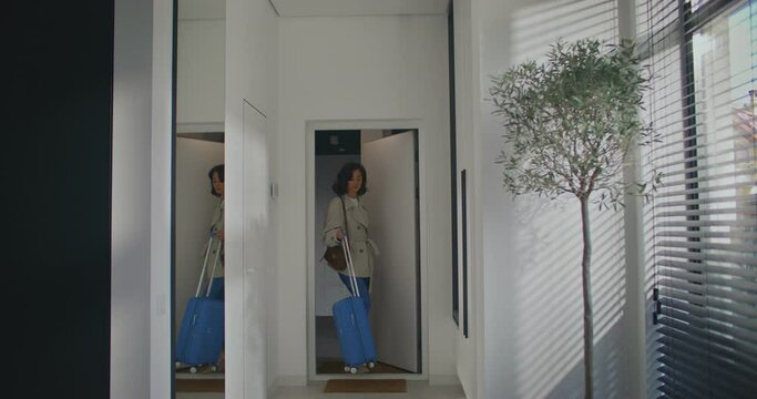 Charming Caucasian girl coming back home from vacation. Leaving blue suitcase on wheels at entrance. Closing door. Afterwards taking out mobile phone from pocket. Entering or typing message.