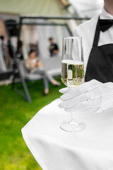 Close-up of a waiter’s hand in white gloves holding a champagne glass, with a blurred background of guests at an outdoor event