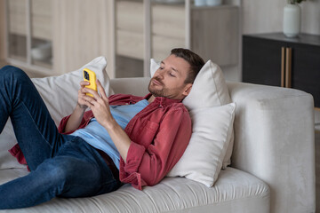 Disinterested man with phone lying on couch surfing online web, procrastination at home. Lazy male...