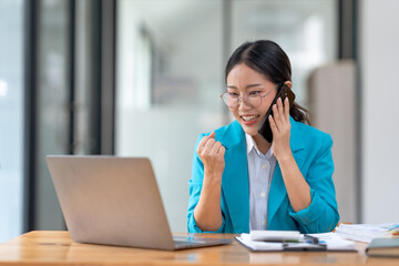 Businesswoman in blue blazer talks on phone and checks laptop at modern office desk, looking focused and smiling at her work.