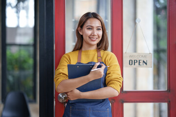 Smiling female barista standing in front of cafe door, holding clipboard, and welcoming customers with a 'Welcome Open' sign.