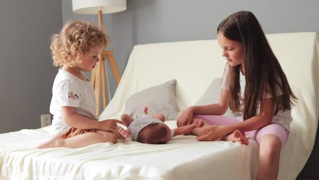 Joyful sisters dressed in comfortable home clothes eagerly gathering on the bed playing gently with their tiny newborn brother scene of pure happiness