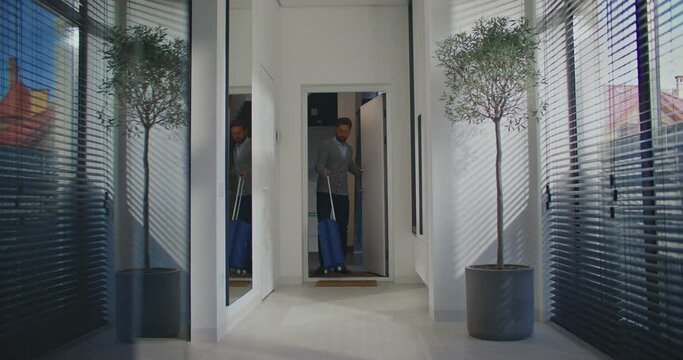 Caucasian male entering apartment. Man using smartphone while dragging small blue suitcase with wheels. Stopping in middle of foyer. Getting distracted by something in next room. Greeting.