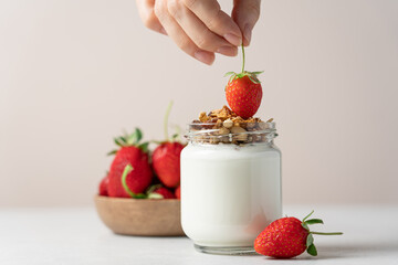 Woman's hand puts strawberries into natural yogurt in a glass jar on a light background with copy...