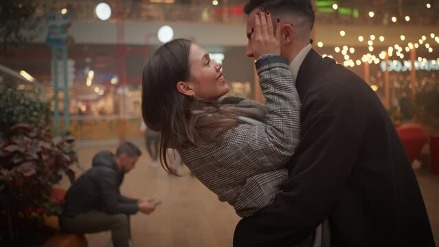 Playful romantic couple rubbing noses and kissing at Christmas market during evening. Side view of cheerful man and woman spending quality time during festive season. 