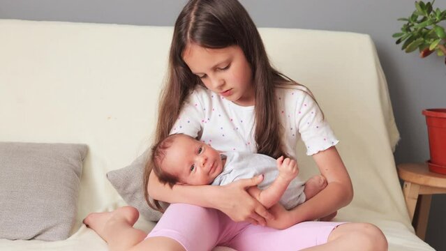 Gentle little girl older sister rocking her little newborn brother on her arms looking at baby with love taking care of baby child while sitting on sofa with in home interior