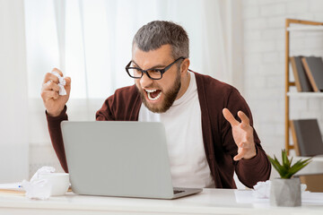 A bearded man with glasses is sitting in a bright office, visibly frustrated and shouting at his...