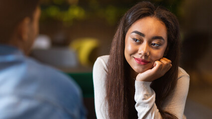 African American girl is sitting at a table in a restaurant, staring intently at a guy who is...