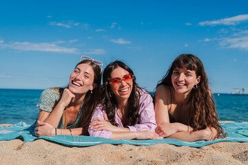 Group of only women lying down on the sand of the beach, smiling and looking at camera. Three young female friends laughing together on the seacoast on a sunny weekend day. Girls enjoying at shore