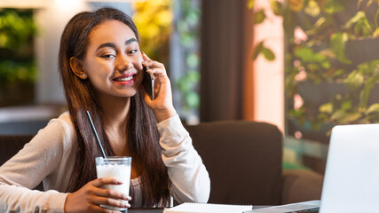 African American young girl is sitting at a table, engaged in a phone conversation. She appears focused on the call, holding the phone to her ear while gesturing with her free hand.