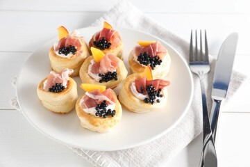 Delicious puff pastry with cream cheese, ham, black caviar, peach and cutlery on white wooden table