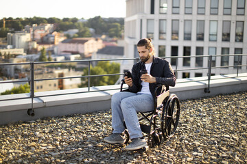 Man in wheelchair using smartphone and drinking coffee on rooftop terrace overlooking cityscape. Casual urban lifestyle.