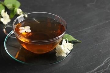 Aromatic jasmine tea in glass cup, flowers and green leaves on grey table, closeup. Space for text