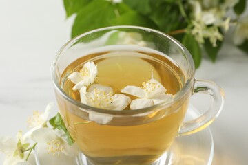 Aromatic jasmine tea in cup, flowers and green leaves on white table, closeup