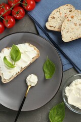 Pieces of bread with cream cheese, basil leaves and tomatoes on gray table, flat lay