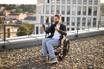 Caucasian man in wheelchair using smartphone for video call on urban rooftop, smiling and engaging in conversation with cityscape behind.