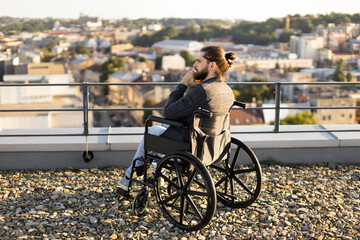 Naklejka premium Thoughtful man in wheelchair observing urban landscape from rooftop, showcasing contemplation and accessibility in urban environments.