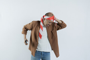 Asian man holding cell phone and showing gesture of disappointment expression. Bald-headed Asian man wearing a headband typical of the independence day of the republic of indonesia