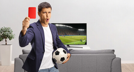 Young man holding a football and showing a red card in front of tv