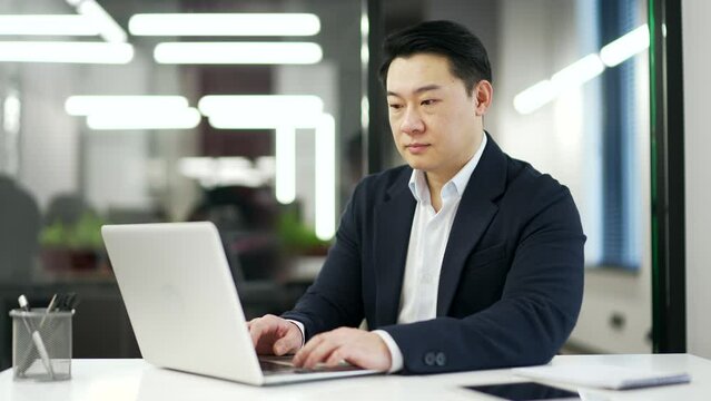 Asian businessman typing on a laptop sitting at a workplace in a business office. Entrepreneur in a formal suit working in computer app, banking, texting a client, chatting online. busy with a project