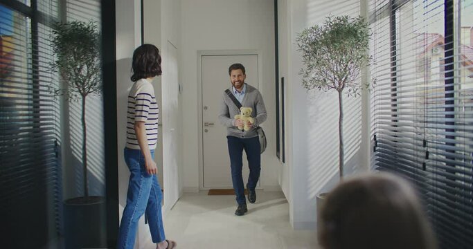 Attractive father coming back home after hard day at work. Busy man not forgetting buying toy for little child. Presenting teddy bear to daughter. Wife coming to greet husband at entrance to house.