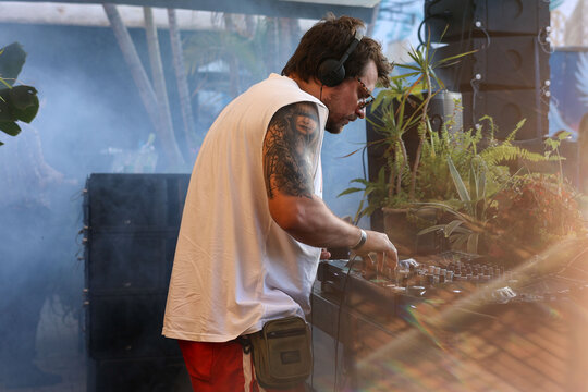 DJ mixing surrounded by plants and smoke wearing casual summer clothes