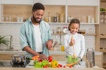 Black father and his daughter give thumbs up while cooking together in the kitchen, enjoying family time and embracing fatherhood