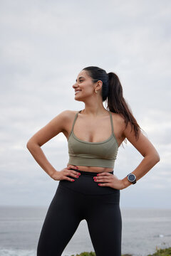 Female hispanic runner posing , arms on hips, big smile, wearing black leggings and a olive top, ocean in the background.