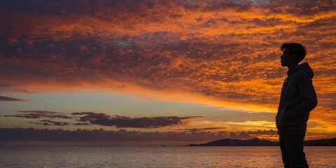 Fototapeta premium A contemplative silhouette stands before a dramatic sunset with fiery clouds and a calm sea
