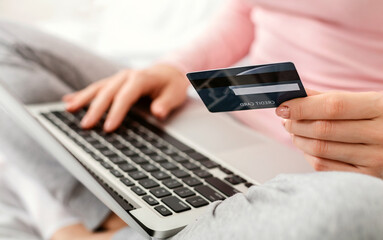 Cropped of woman sits at a desk, holding a credit card in one hand while typing on a laptop with the other hand, focused on entering information into the computer, shopping online