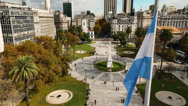 Dozens of people walk through Plaza de Mayo. Steady aerial camera with Argentine flag in foreground