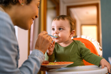 Loving mother spoon-feeding her happy baby boy. Adorable toddler in green outfit enjoying a...