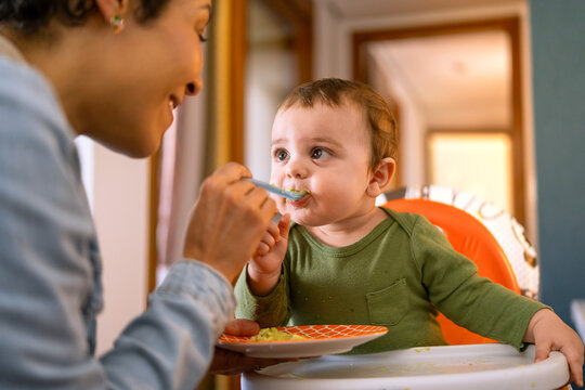 Loving mother spoon-feeding her happy baby boy. Adorable toddler in green outfit enjoying a nutritious meal at home..