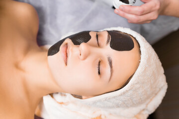 Cosmetologist applying black sheet mask on woman's face in beauty salon.