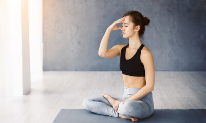 A young woman is seated in the lotus position, practicing breathing exercises with her eyes closed. She is indoors on a yoga mat, wearing a black sports top and gray leggings, sun flare